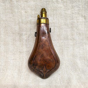 Könnte beinhalten: Antike Pulverflasche aus Kupfer mit Messingakzenten. Die Flasche hat eine tropfenförmige Form mit einem dekorativen Blumendesign an der Basis. Die Oberseite hat eine Messingdüse und einen kleinen Ring zum Aufhängen.