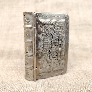 Puede incluir: Un pequeño objeto metálico de tono plateado con forma de libro en miniatura. El lomo presenta bandas horizontales en relieve con patrones estampados, mientras que la cubierta frontal muestra un intrincado diseño geométrico y floral en relieve.