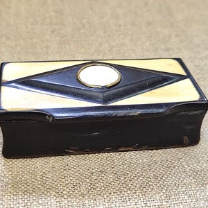 Puede incluir: Una caja rectangular con base de color marrón oscuro y una tapa con incrustaciones de color crema. La tapa muestra una forma de diamante de color marrón oscuro centrada con una incrustación circular crema rodeada por un anillo de metal dorado.