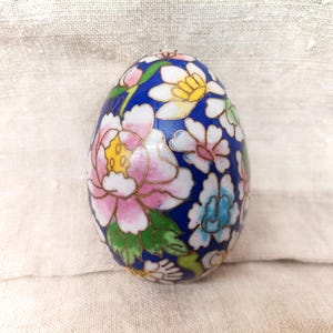Op de afbeelding: Een blauw, eivormig sieraad met een bloemenmotief. Het ei is versierd met roze, witte en blauwe bloemen op een donkerblauwe achtergrond.