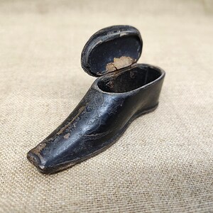 Puede incluir: Un pequeño recipiente negro con forma de zapato y tapa abatible. La superficie exterior muestra signos de desgaste y textura, con un acabado oscuro y desgastado. La tapa está abierta, revelando el interior hueco del objeto en forma de punta.