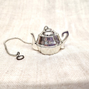 Può includere: Infusore per tè color argento a forma di teiera, con catena e anello. L'infusore ha un coperchio con piccoli fori e un manico. Lo sfondo è un tessuto strutturato bianco sporco. Per tè sfuso.