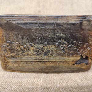 Puede incluir: Una placa rectangular de madera de color marrón oscuro con un relieve tallado de La Última Cena. La escena muestra figuras sentadas a una mesa larga con detalles arquitectónicos de fondo. La madera presenta vetas naturales.