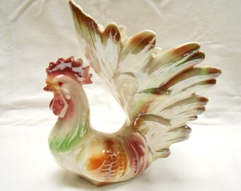 Peut inclure: Figurine de coq en céramique avec un design détaillé. Le coq a une crête et un fanon rouges, avec un corps aux nuances d'orange, de vert et de marron. Les plumes de la queue sont grandes et s'évasent, avec une finition nacrée.