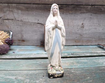 Puede incluir: Una estatua de yeso blanco de la Virgen María, de pie sobre una base negra. Lleva una túnica blanca con una faja azul y un velo blanco. Tiene las manos juntas en oración.