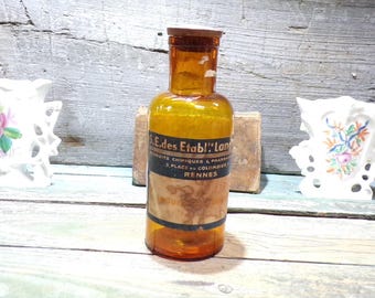 May include: An amber glass bottle with a wooden stopper. The bottle has a label with French text. The bottle is cylindrical with a rounded shoulder and a narrow neck. The label is dark with light text. The bottle is likely an antique.