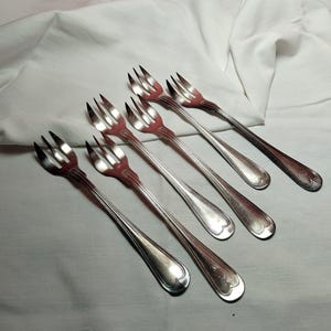 May include: Six silver-colored forks with three tines each are arranged diagonally on a white cloth. The forks have a simple, elegant design with a rounded end. The tines are slightly curved.
