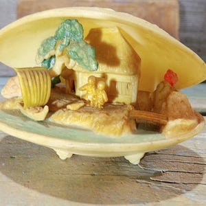 Könnte beinhalten: Dekorative Muschelschalenfigur mit einer Miniatur-Dorfszene. Die Szene umfasst ein kleines Haus, eine Brücke und Figuren in Gelb-, Braun- und Grüntönen. Die Muschel liegt auf einer Holzoberfläche.
