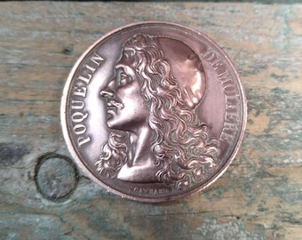 Puede incluir: Medallón redondo de color bronce con el perfil de un hombre con pelo largo y rizado. Las palabras "POQUELIN" y "DE MOLIERE" están inscritas en el borde. El medallón está sobre una superficie de madera envejecida.