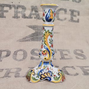 May include: A white ceramic candlestick with colorful floral and leaf designs in red, yellow, and blue. The candlestick has a flared base and a cup-shaped top, and stands approximately 8 inches tall.
