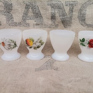 Può includere: Quattro portauova bianchi con disegni floreali. Una tazza presenta una margherita, un'altra una pesca e una terza una rosa rossa. La quarta tazza è semplice. Questi portauova vintage sono perfetti per la tavola della colazione.
