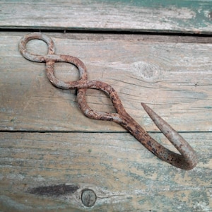 Op de afbeelding: Een roestige metalen haak met een decoratieve lusvormige handgreep. De haak is gemaakt van ijzer en heeft een puntig uiteinde. De haak ligt op een verweerde houten ondergrond.