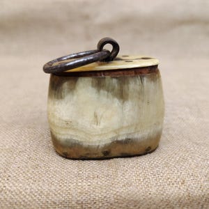 Puede incluir: Un pequeño recipiente de forma ovalada hecho de cuerno natural con un acabado moteado en crema y marrón. Cuenta con una tapa plana de color claro asegurada con un asa de anillo de metal oscuro en la parte superior y pequeños remaches.
