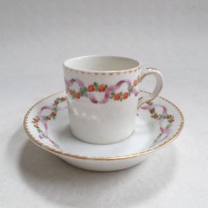 Puede incluir: Un juego de taza y platillo de porcelana blanca con un diseño floral rosa y naranja. La taza y el platillo tienen un borde dorado.