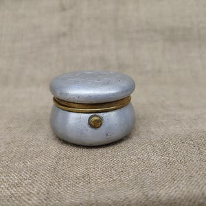 May include: A small, round, silver-coloured metal container with a lid. The container features a brass-coloured rim where the lid meets the base and a small, circular brass-coloured button or latch mechanism on the front side of the lower section.