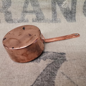 May include: A small copper saucepan with a long handle. The pot has a rounded body and a handle attached with a rivet. The copper has a warm, reddish-brown colour and shows signs of age. The handle is approximately 13 cm long.