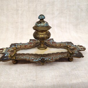 Op de afbeelding: Antieke messing inktpot met een decoratieve lade. De inktpot heeft een deksel met blauwe en witte emaille accenten. De lade heeft sierlijke details met rode en blauwe emaille en staat op kleine messing voetjes. Een vintage bureau-accessoire.