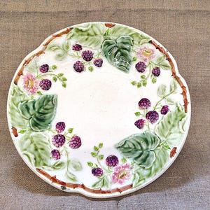 May include: A round ceramic plate featuring a raised, embossed design of dark purple blackberries, light green leaves, and small pink flowers. The rim of the plate is textured to resemble brown thorny bramble branches against a white background.