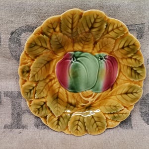 May include: A decorative ceramic plate with a leaf and fruit design. The plate features a central image of two apples, one green and one red, surrounded by detailed yellow-green leaves. The plate has a scalloped edge.