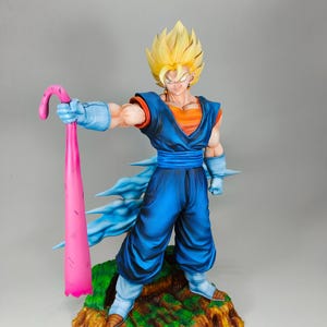 May include: A detailed figurine of a character with spiky yellow hair, wearing a blue and orange outfit. The figure is holding a pink, curved object and stands on a textured, green and brown base. The figure has a determined expression.