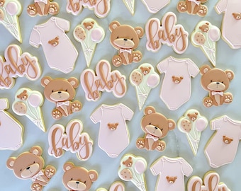 May include: A collection of decorated sugar cookies featuring a baby theme. The cookies are decorated with pink and brown icing and feature various designs, including bears, baby onesies, and the word "baby".