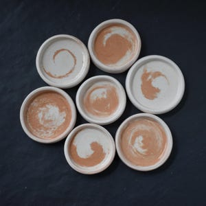 May include: A collection of seven small, round ceramic dishes. Each dish has a white exterior and a peach-colored swirl design in the center. The dishes are arranged on a dark surface.