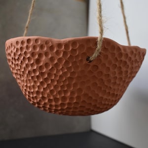 May include: A terracotta-colored hanging bowl with a textured surface featuring numerous small, circular indentations. The bowl has an uneven, wavy rim and is suspended by three strands of natural jute twine threaded through holes in the side.
