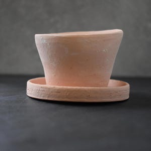 May include: A light peach-colored terracotta plant pot with a matching circular saucer. The pot features a wide, slightly angled rim and a smooth, matte finish, while the saucer has a textured, rustic edge and a flat, circular base design.