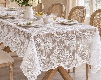 May include: A rectangular white lace tablecloth featuring an intricate floral pattern with scalloped edges, draped over a wooden dining table. The tablecloth is semi-sheer, revealing the wood surface underneath, and is styled with white plates.