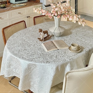 Puede incluir: Un mantel redondo blanco con un detallado patrón de cachemira gris sobre una mesa de comedor. En la mesa hay un jarrón blanco con flores rosas, una figura de caballo balancín de madera, un libro abierto y una taza Café de France.