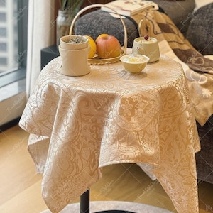 May include: A round table covered with a beige tablecloth featuring a tonal damask pattern. On the table sits a beige ceramic warmer, a woven basket containing a red apple and a yellow fruit, a small beige teapot, and a small beige bowl.