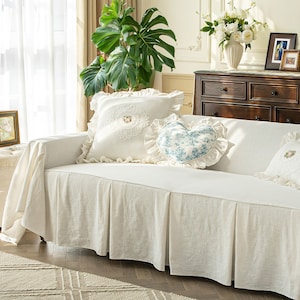 Puede incluir: Una funda de sofá de lino blanco con una falda con volantes y una almohada decorativa en forma de corazón. El sofá está decorado con dos almohadas adicionales, una con un diseño floral y la otra con un borde de encaje.