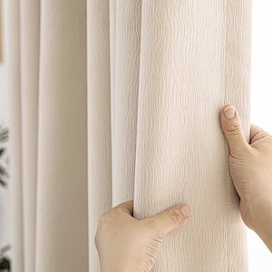 May include: Close-up of a cream-colored curtain with a textured weave. The curtain is being held open by a hand.