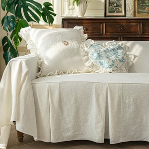 May include: A white linen sofa slipcover with a ruffled edge and a pleated skirt. The sofa is decorated with two white pillows, one with a floral design and the other with a heart-shaped design.