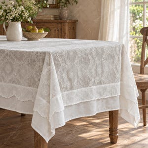 May include: A rectangular beige tablecloth featuring intricate white lace embroidery with floral and geometric patterns. The fabric has a layered appearance with a scalloped edge border, draped over a wooden table surface with a visible texture.