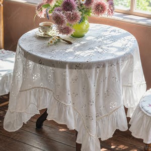 Könnte beinhalten: Eine runde weiße Tischdecke mit einem komplizierten floralen Spitzenmuster und einem gerüschten Saum. Der Stoff weist durchgehend ausgeschnittene Details auf. Die Tischdecke liegt auf einem runden Tisch und erzeugt durch die gerafften Kanten eine weiche Optik.