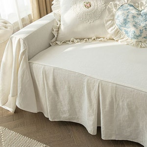 May include: A white linen sofa slipcover with a ruffled edge and a pleated skirt. The slipcover is on a sofa with two pillows, one with a floral design and the other with a heart shape.