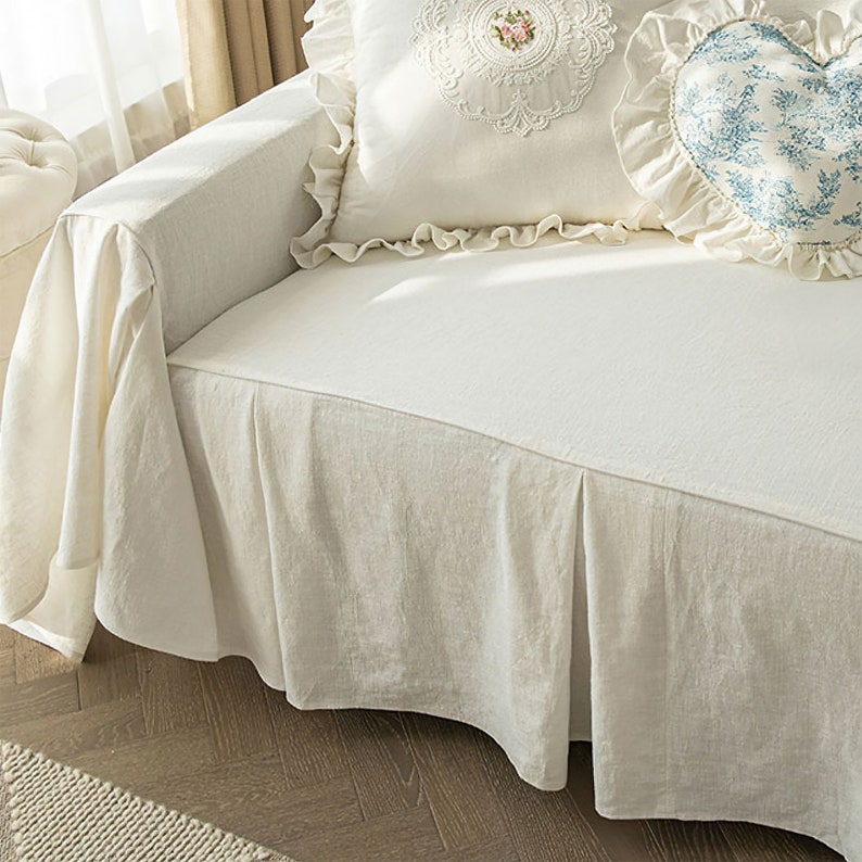 May include: A white linen sofa slipcover with a ruffled edge and a pleated skirt. The slipcover is on a sofa with two pillows, one with a floral design and the other with a heart shape.
