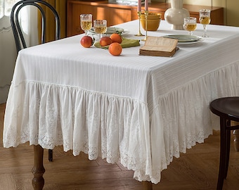 May include: A white rectangular tablecloth featuring a subtle vertical striped texture and a wide, gathered ruffled skirt along the edges. The bottom hem of the ruffle is finished with an intricate, delicate white floral lace trim detail.