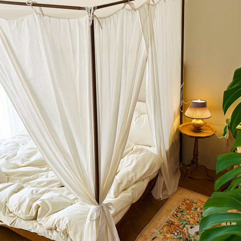 May include: A white canopy bed with white bedding and a wooden frame. A small table with a lamp sits next to the bed.