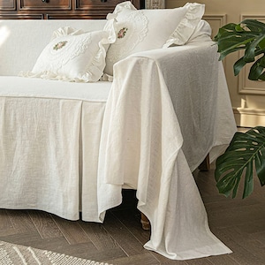 May include: A white linen sofa slipcover with a ruffled edge and a white lace pillow with floral embroidery. The slipcover has a gathered skirt and is draped over the back of the sofa.