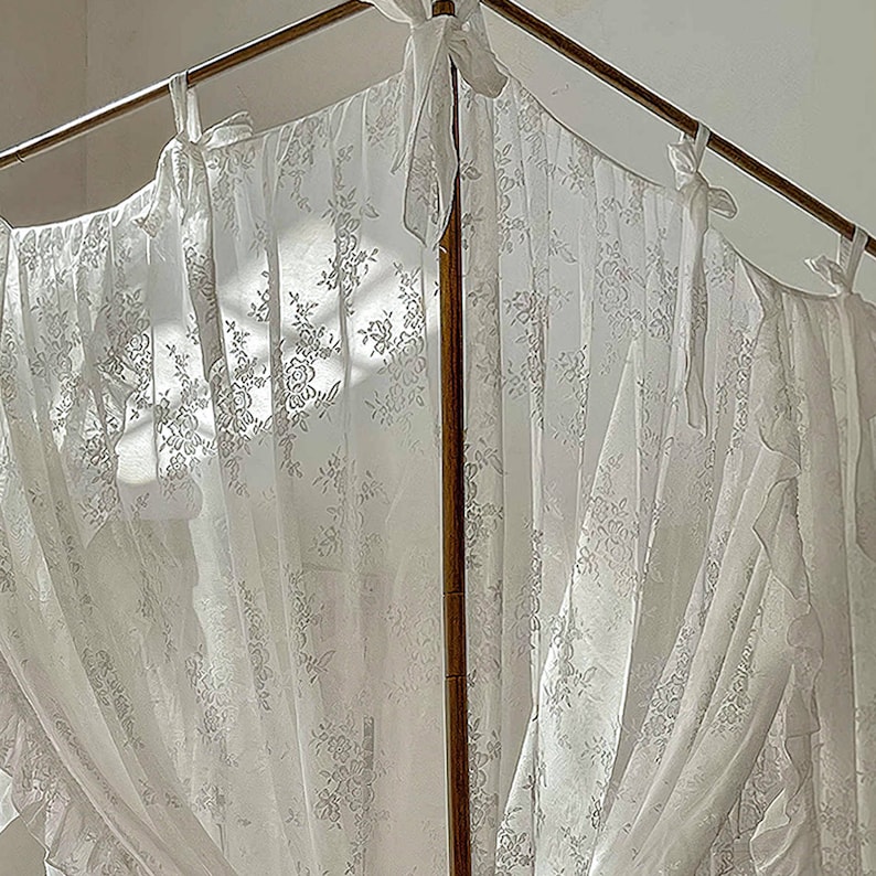 May include: White lace curtains with a floral pattern hang from a wooden rod. The curtains are gathered at the top and bottom, creating a soft and romantic look.