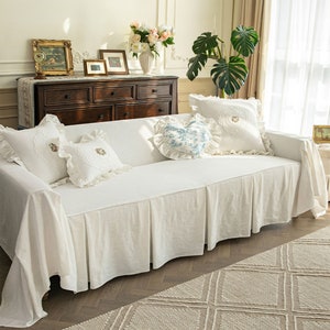 May include: White sofa slipcover with a ruffled skirt and embroidered pillows. The slipcover is made of a soft, linen-like fabric and features a simple, elegant design.