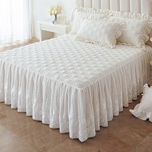 May include: White bed skirt with a lace trim and a quilted design. The bed skirt is made of a soft, luxurious fabric and is perfect for adding a touch of elegance to any bedroom.