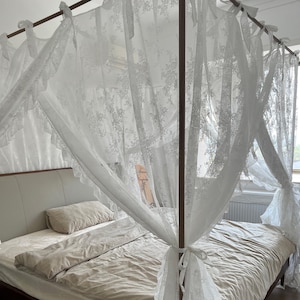 May include: A white lace canopy bed with a beige duvet and two pillows. The canopy is draped over the bed frame and tied at the corners.