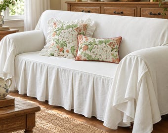 May include: A white sofa cover with a pleated skirt and ruffled edges, featuring two decorative throw pillows. The square pillow has a ruffled border, while the rectangular pillow has a red piped edge. Both pillows display a floral pattern.
