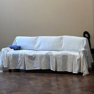 May include: A white sofa covered with a white sheet, with a blue pillow on the seat. The sofa is in a room with a wooden floor.