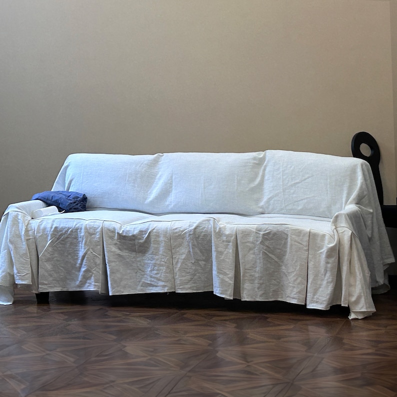 May include: A white sofa covered with a white sheet, with a blue pillow on the seat. The sofa is in a room with a wooden floor.