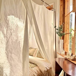 May include: A white canopy bed with a white bedspread and pillows. The bed is in a room with a window and a wooden table.