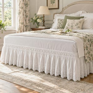 May include: A white bed skirt featuring a delicate eyelet lace trim along the bottom edge. The fabric is solid white with a gathered, ruffled design that hangs from the base of the mattress to the floor, providing a decorative finish to the bed.